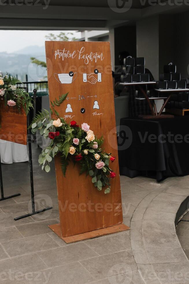 A front board sign translate in Indonesia say Health protocol, with flower decoration standing at the wedding entrance. photo