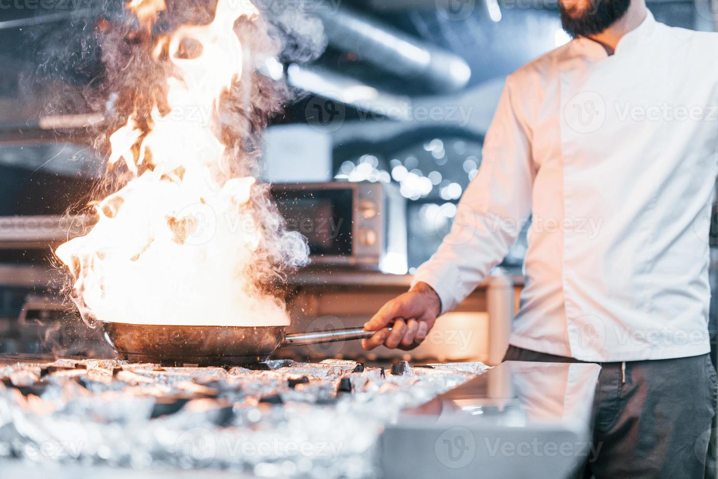 Frying pan is on fire. Chef in white uniform cooking food at kitchen. Busy day at work photo