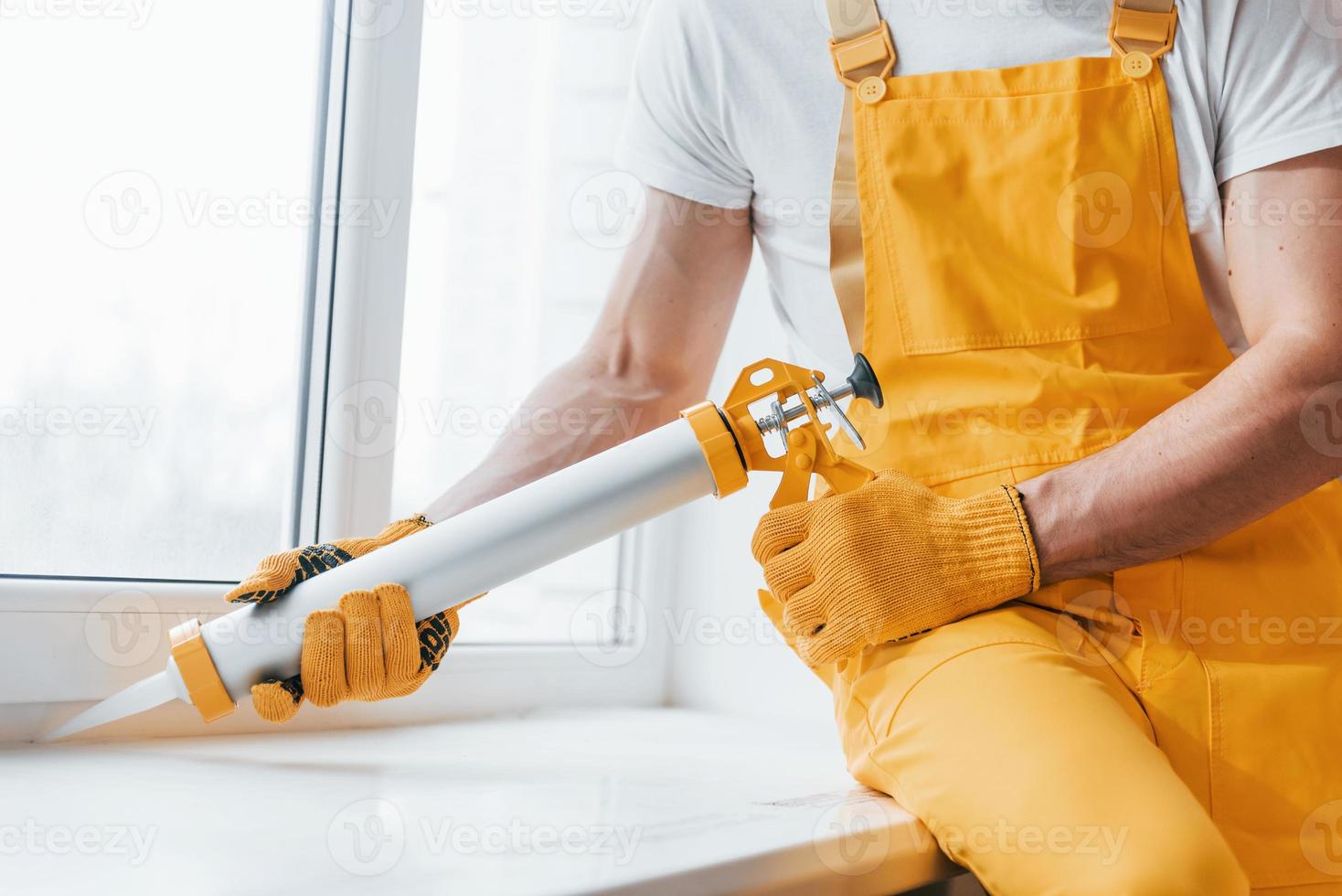 Handyman in yellow uniform works with glue for window indoors. House renovation conception photo