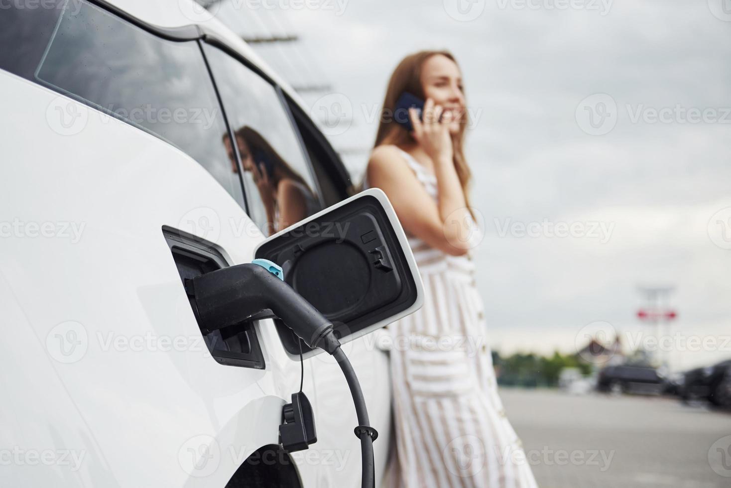 Having conversation by using phone. Woman on the electric cars charge station at daytime. Brand new vehicle photo