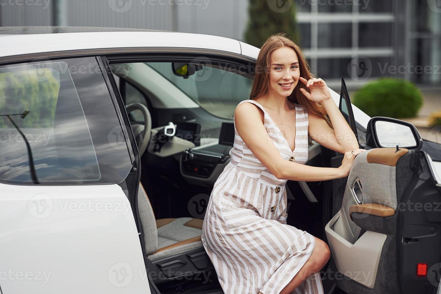 Smiling blonde sits in the white car. Business building behind photo