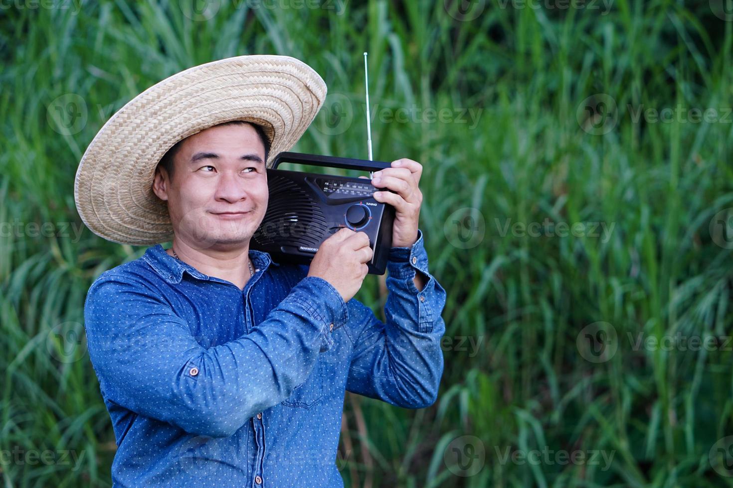 Asian man farmer wears hat, blue shirt, holds transistor radio on his shoulder at garden, feels happy. Concept , Happy working along with music, news, information  from radio. Country lifestyle. photo