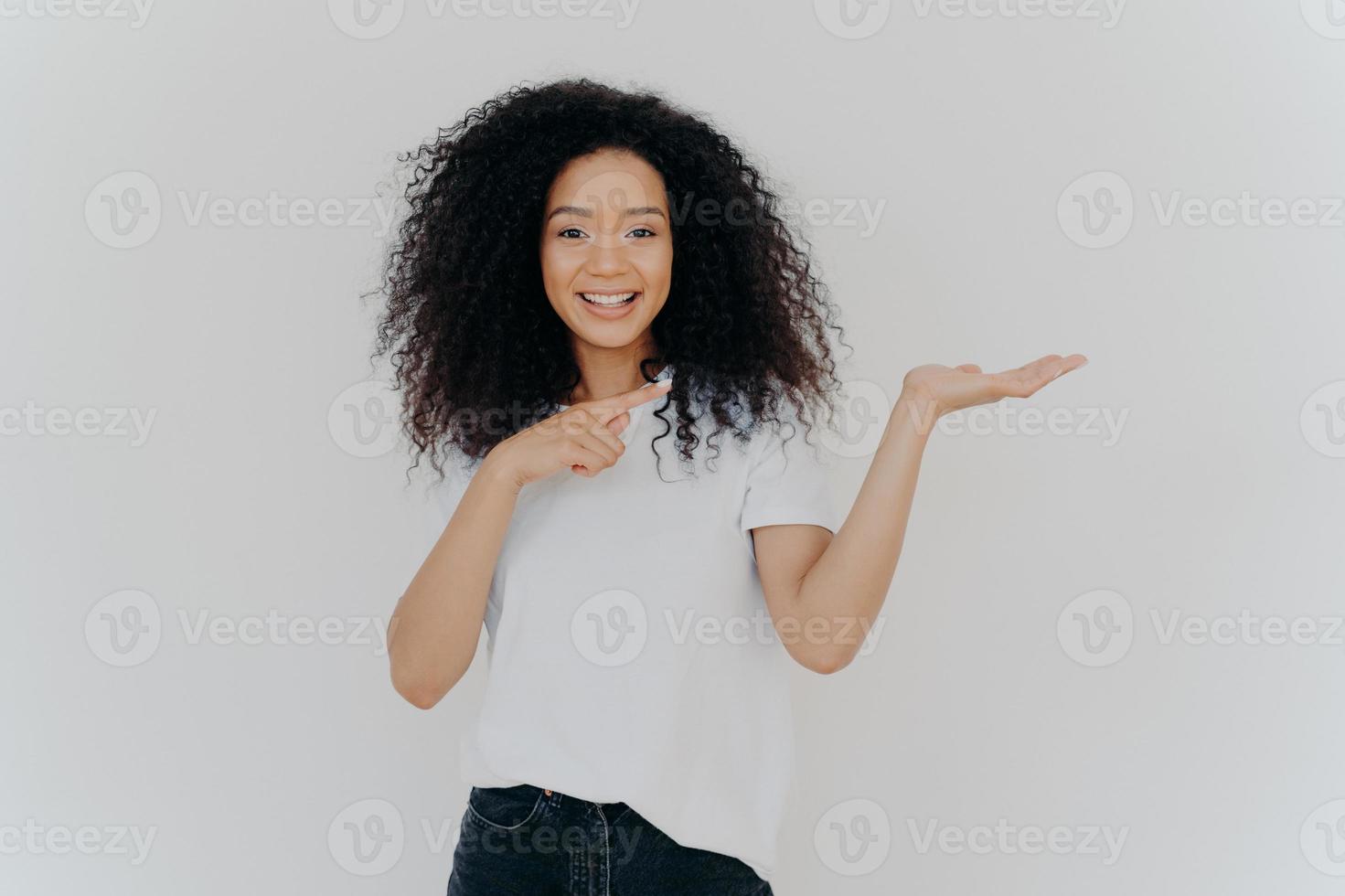 Choice is simple. Attractive feminine girl with Afro hair raises palm and points on blank space, makes decision, has toothy smile, wears casual wear, poses against white background. Look at this photo