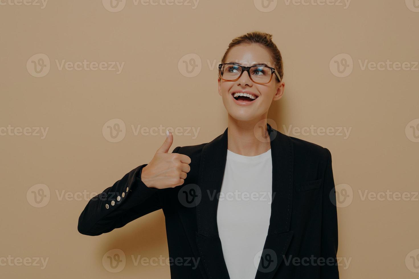 Portrait of satisfied female in eyeglasses smiling while showing thumb up with hand photo