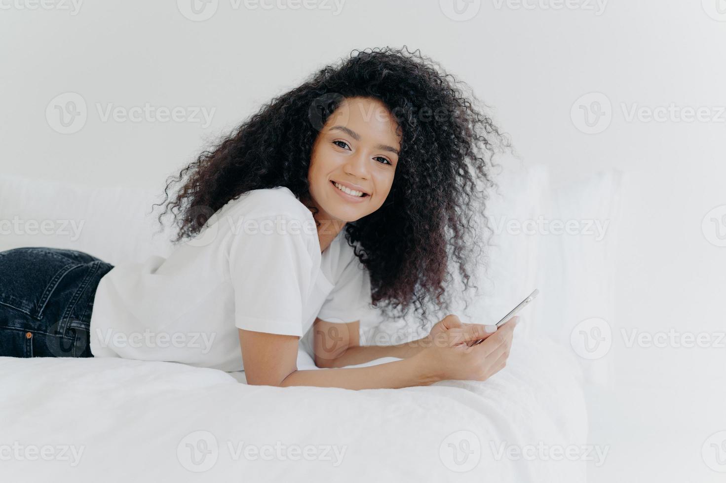 Pretty cheerful woman with Afro haircut, enjoys chatting and networking, lies on comfortable bed, looks photos, has relaxed face expression, isolated over white background. Online communication photo