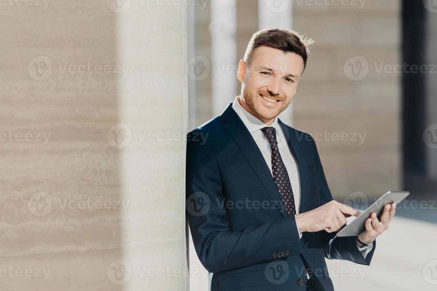Experienced male economist in formal clothes works on digital tablet, has work break, stands in office interior with copy space. Successful entrepreneur prepares business report on modern device photo
