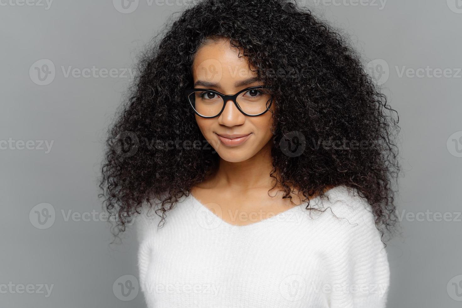 Close up shot of dark skinned female has satisfied expression, bushy curly hair, wears spectacles and white jumper, looks straightly at camera, models over grey background. Natural beauty concept photo