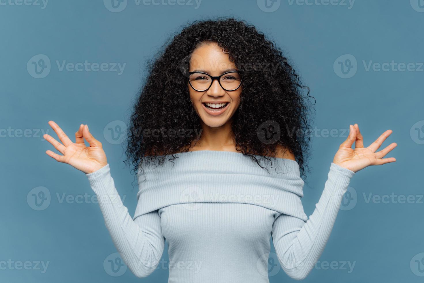 una dama afroamericana positiva hace un buen gesto con ambas manos, medita en el interior, usa suéter y anteojos de moda, está de buen humor, aislada sobre un fondo azul. concepto de lenguaje corporal foto