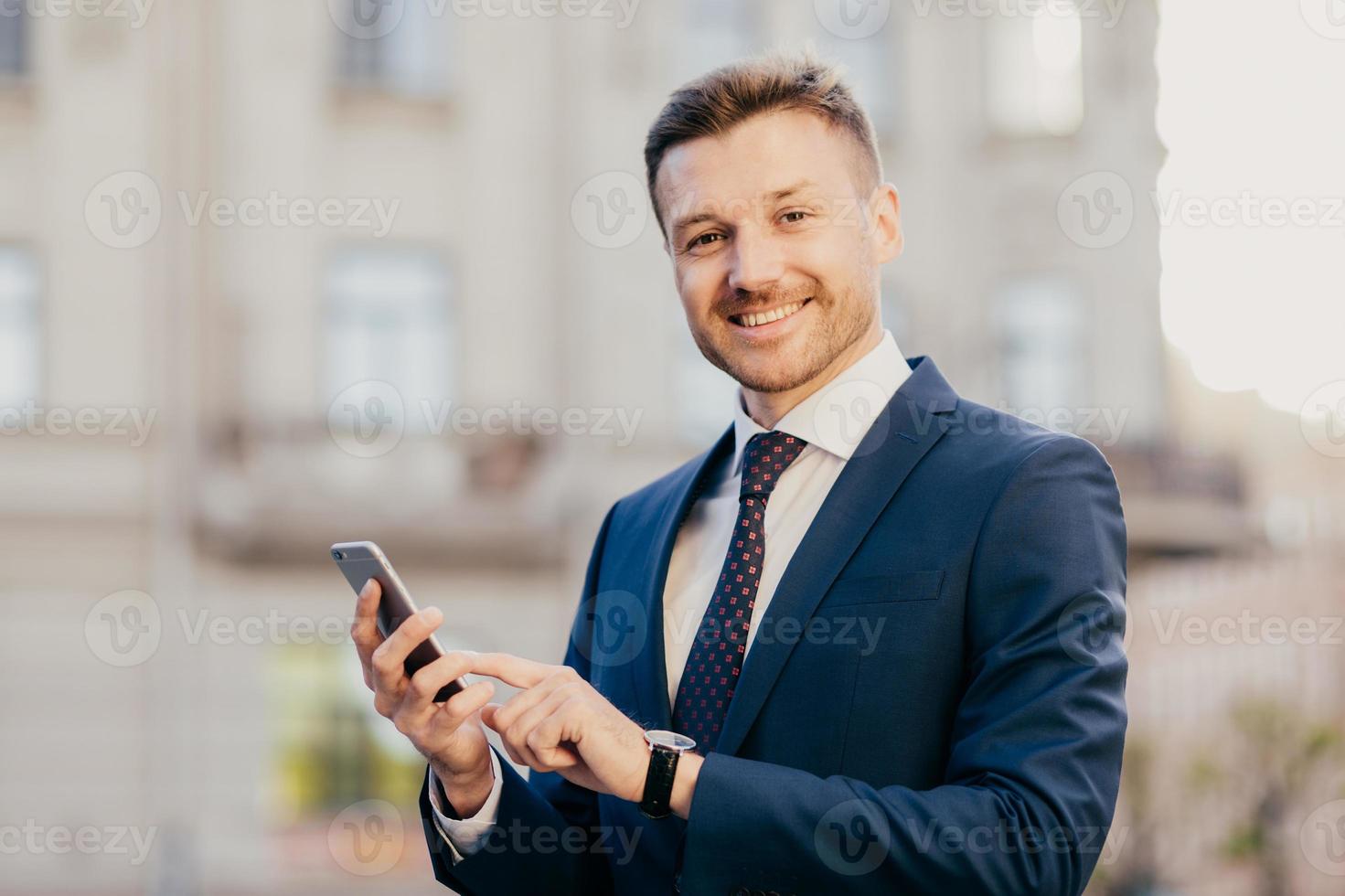 Cheerful male financier holds modern cell phone chats with partners, arrange business meeting, poses outdoors, has happy expression, dressed in black elegant suit. Successful businessman reads message photo