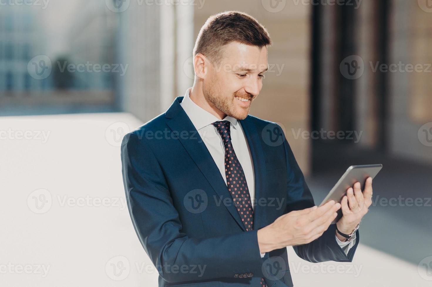Cheerful male manager reads notification on digital tablet, wears formal black suit with tie, connected to wireless internet, watches video on web page. Business and modern technologies concept photo