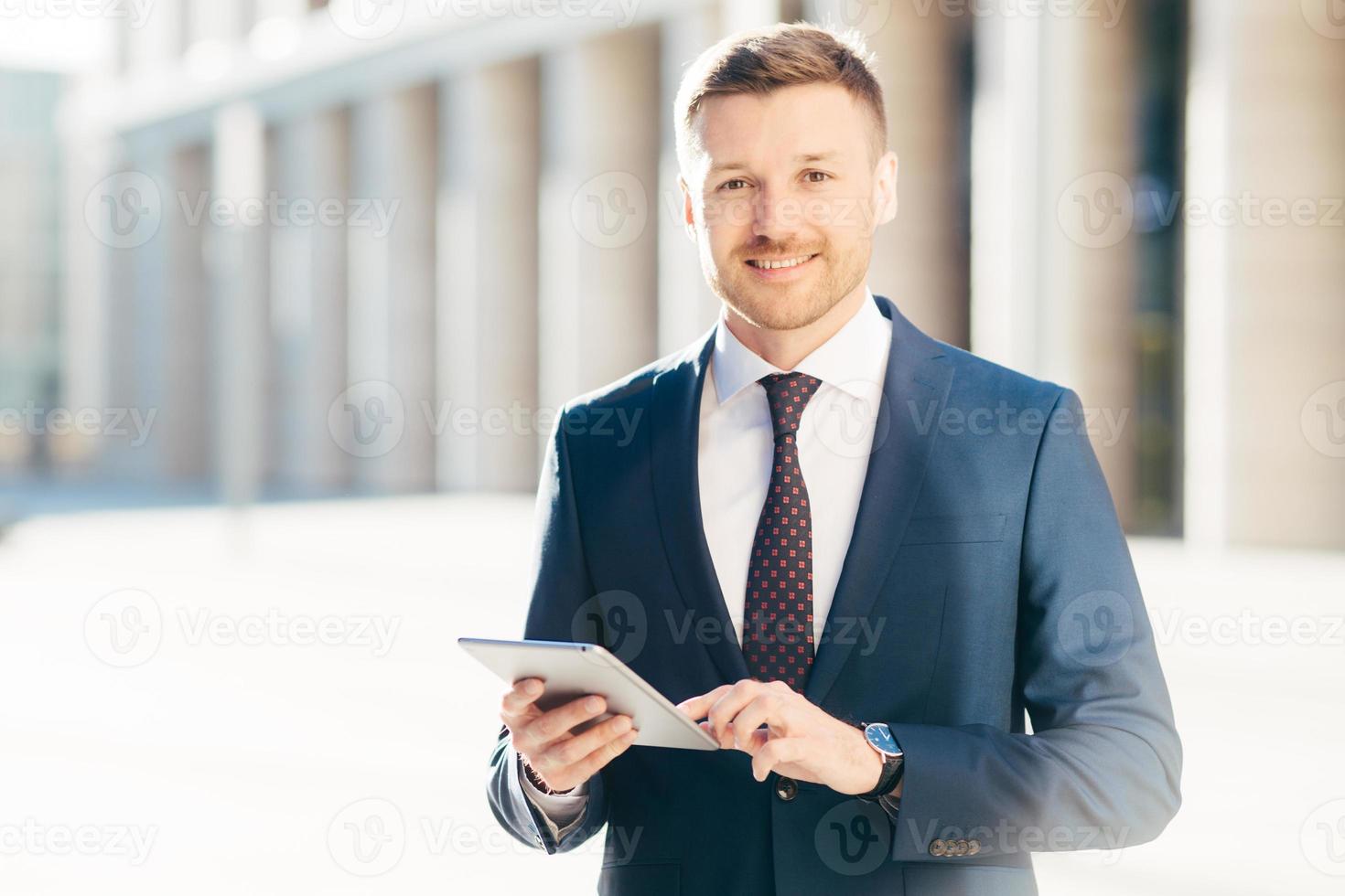 Cheerful successful young male executive manager, wears formal clothes, checks email box on modern touchpad, uses modern device for online banking app, has pleasant appearance. Business concept photo