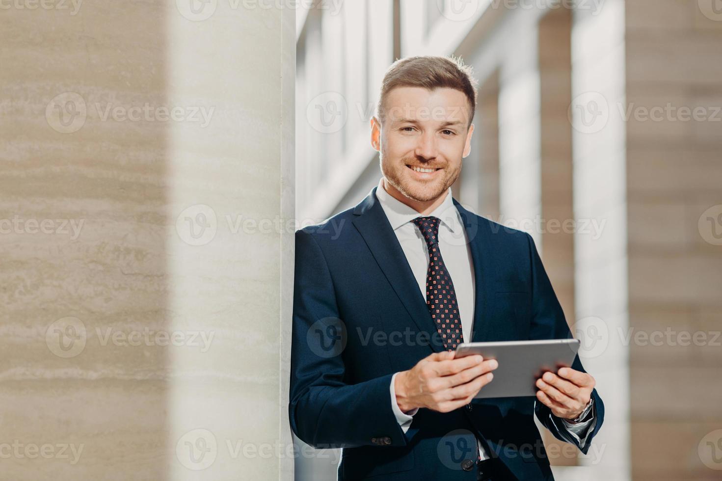 Confident cheerful male lawyer reads business news, has gentle smile, dressed in formal clothes, poses in urban setting. Businessman checks email or updates profile on digital tablet computer photo