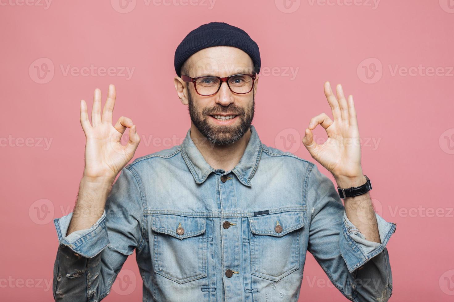 Cheerful bearded male in fashionable clothing, makes ok sign, being satisfied with something, isolated over pink studio background. Unshaven middle aged man gestures indoor, shows his approval photo