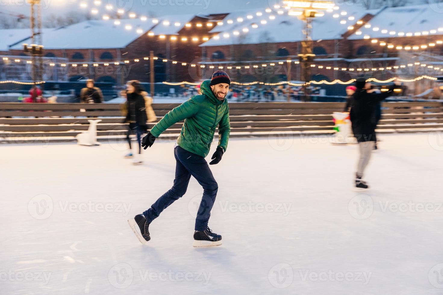 Joyful bearded man practices going skating on ice ring, has cheerful expression, smiles happily, demontrates his professionalism. Active sporty male in green jacket go in for winter sport photo