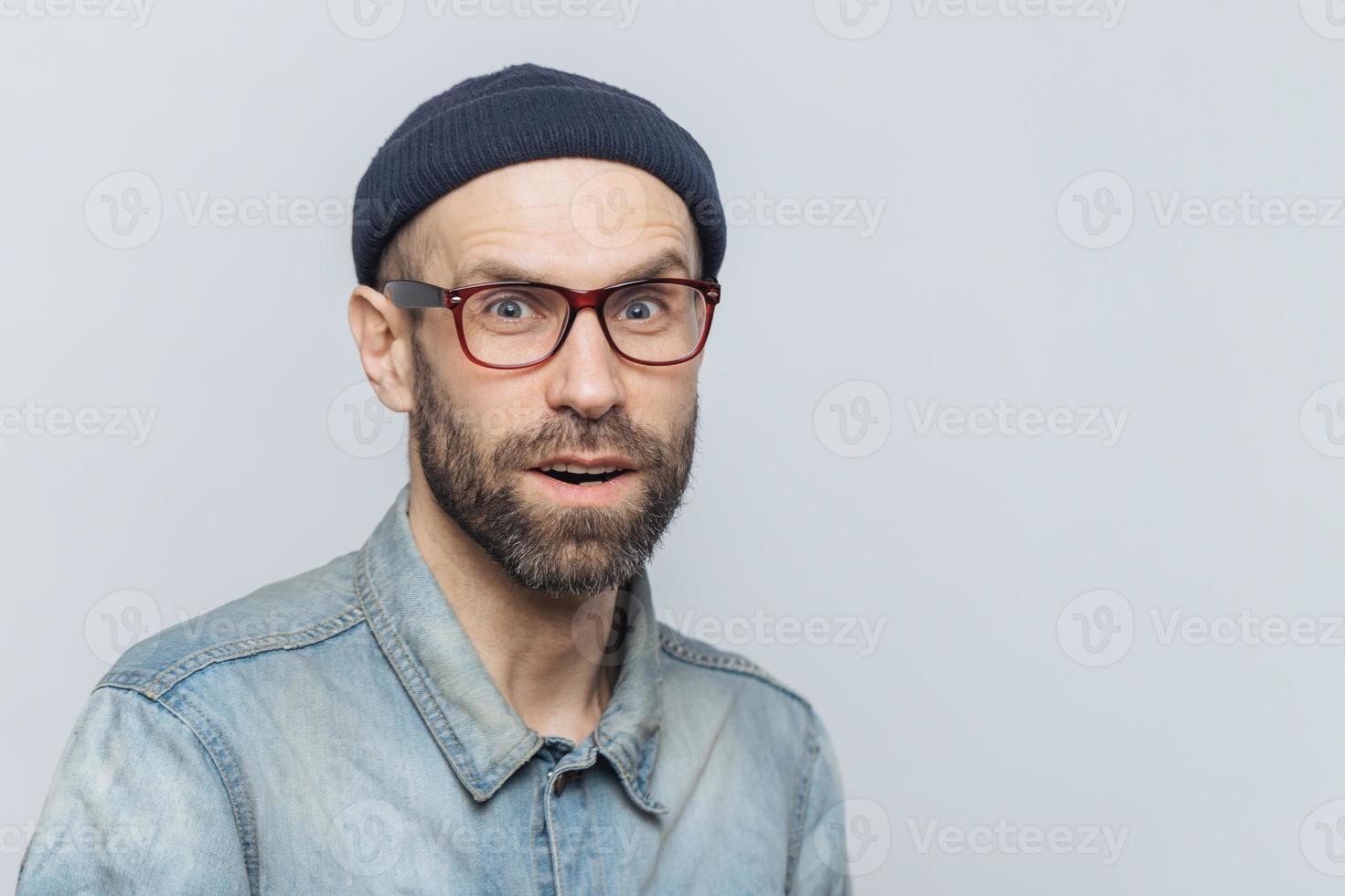 Stylish middle aged male with suprised and curious expression wears spectacles, denim jacket and hat, poses against grey background with copy space for your advertisment or promotional text. photo