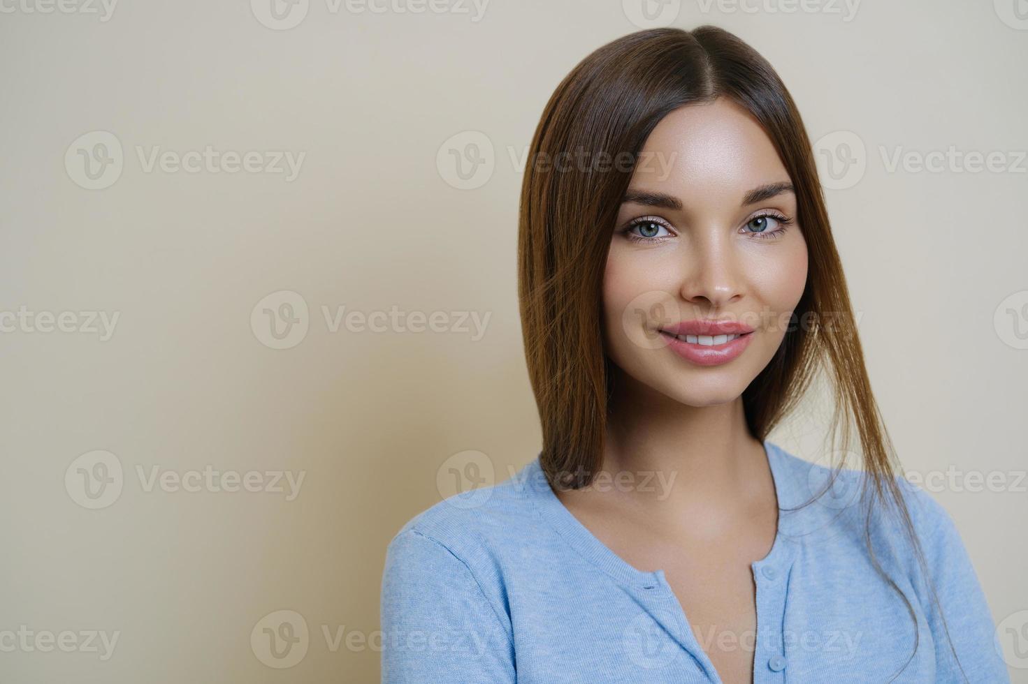 Portrait of lovely dark haired woman with pleasant appearance, full lips, wears casual blue jumper, poses against beige background, has calm expression sensual look healthy skin after facial treatment photo