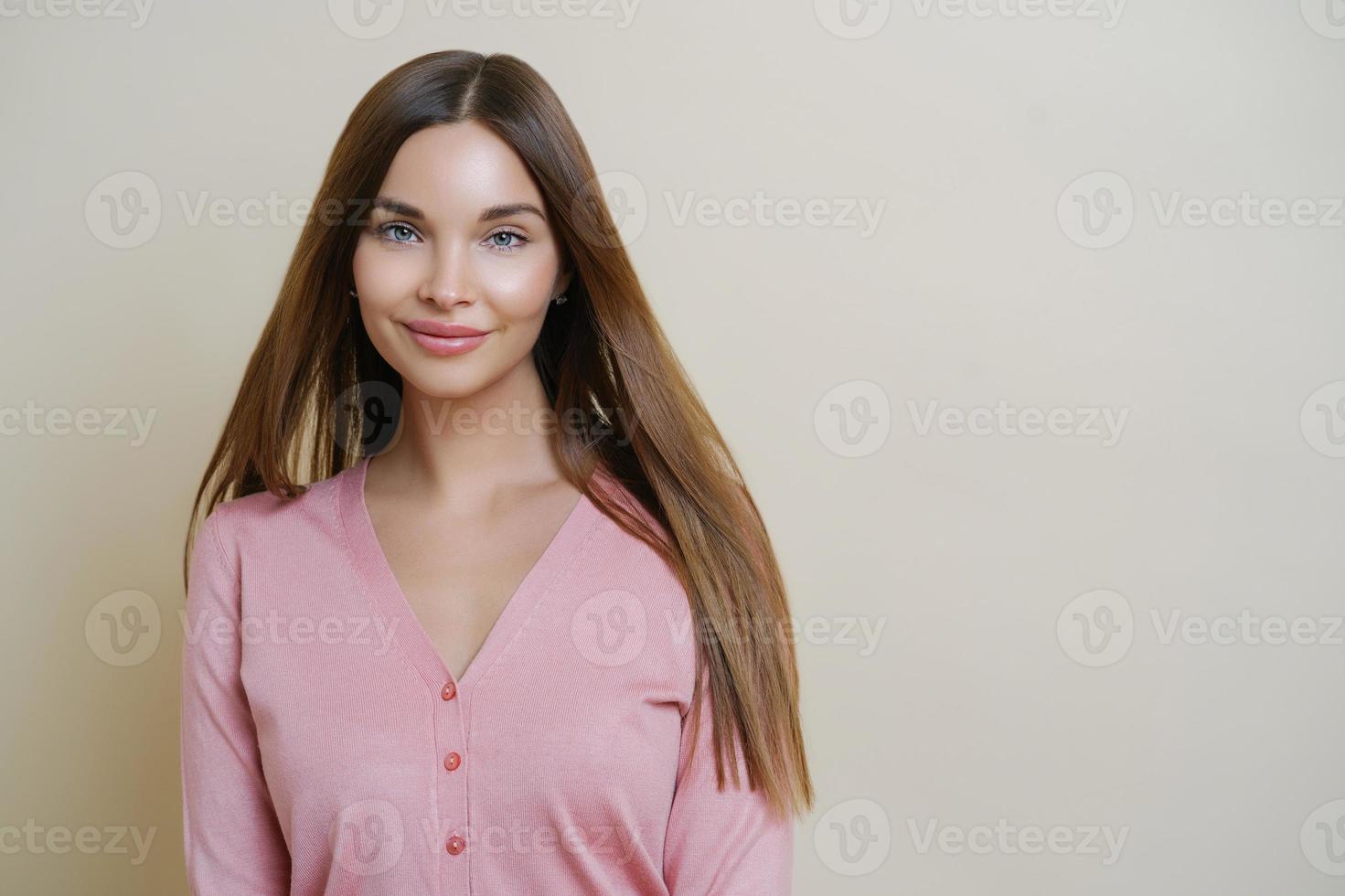 el retrato horizontal de una encantadora mujer europea sonríe suavemente a la cámara, tiene el pelo largo y liso, usa un suéter rosado casual, va a caminar al aire libre, se para contra el fondo beige del estudio. foto