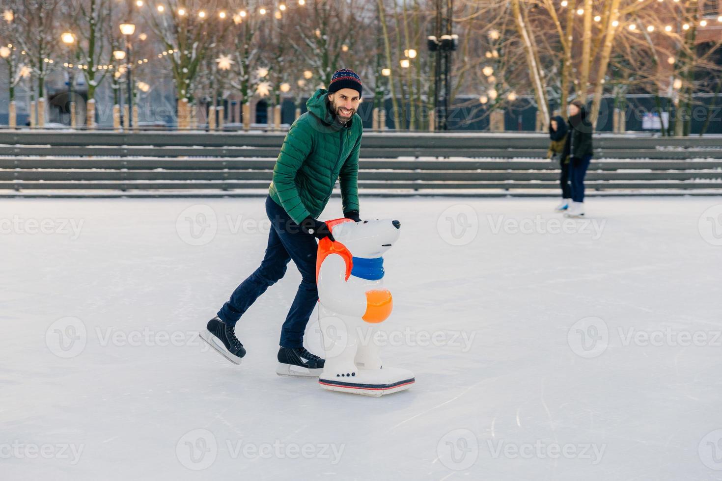 Positive smiling attractive man in warm winter clothes and skates, practices go skating on ice, uses skate rink, looks delightfully into camera, spends Sunday outdoor during winter period photo