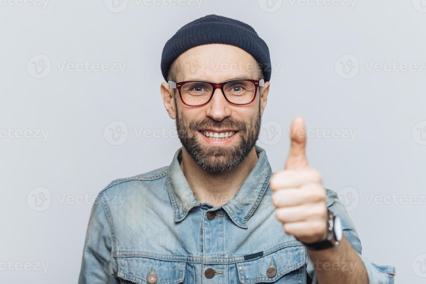 Positive middle aged male with thick beard and mustache, shows ok sign, raises thumb, dressed in fashionable clothing, isolated over white background. People, body language, emotions concept photo