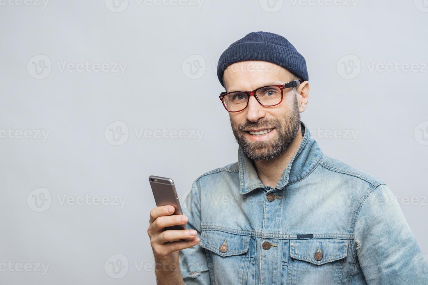 Stusio shot of happy bearded man with stubble holds modern smart phone in hands, enjoys online communication, connected to wireless internet, isolated over white background. Communication concept photo