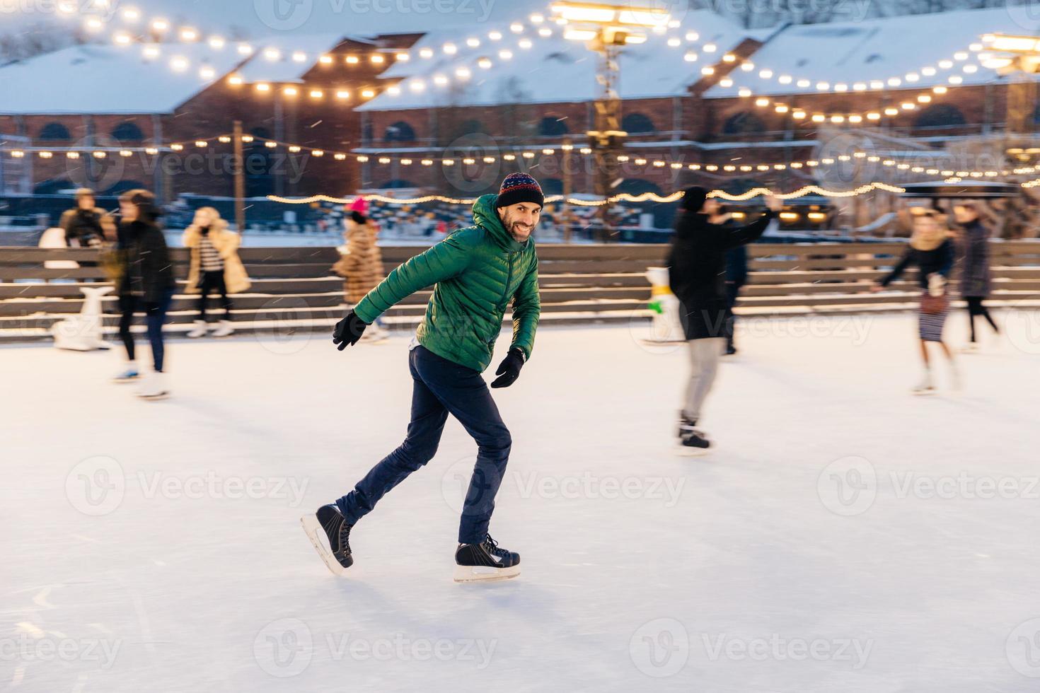 Outdoor shot of attractive man with beard, wears warm winter clothes, practices going skating on ice skate ring decorated with lights, has glad expression, enjoys his favourite action photo