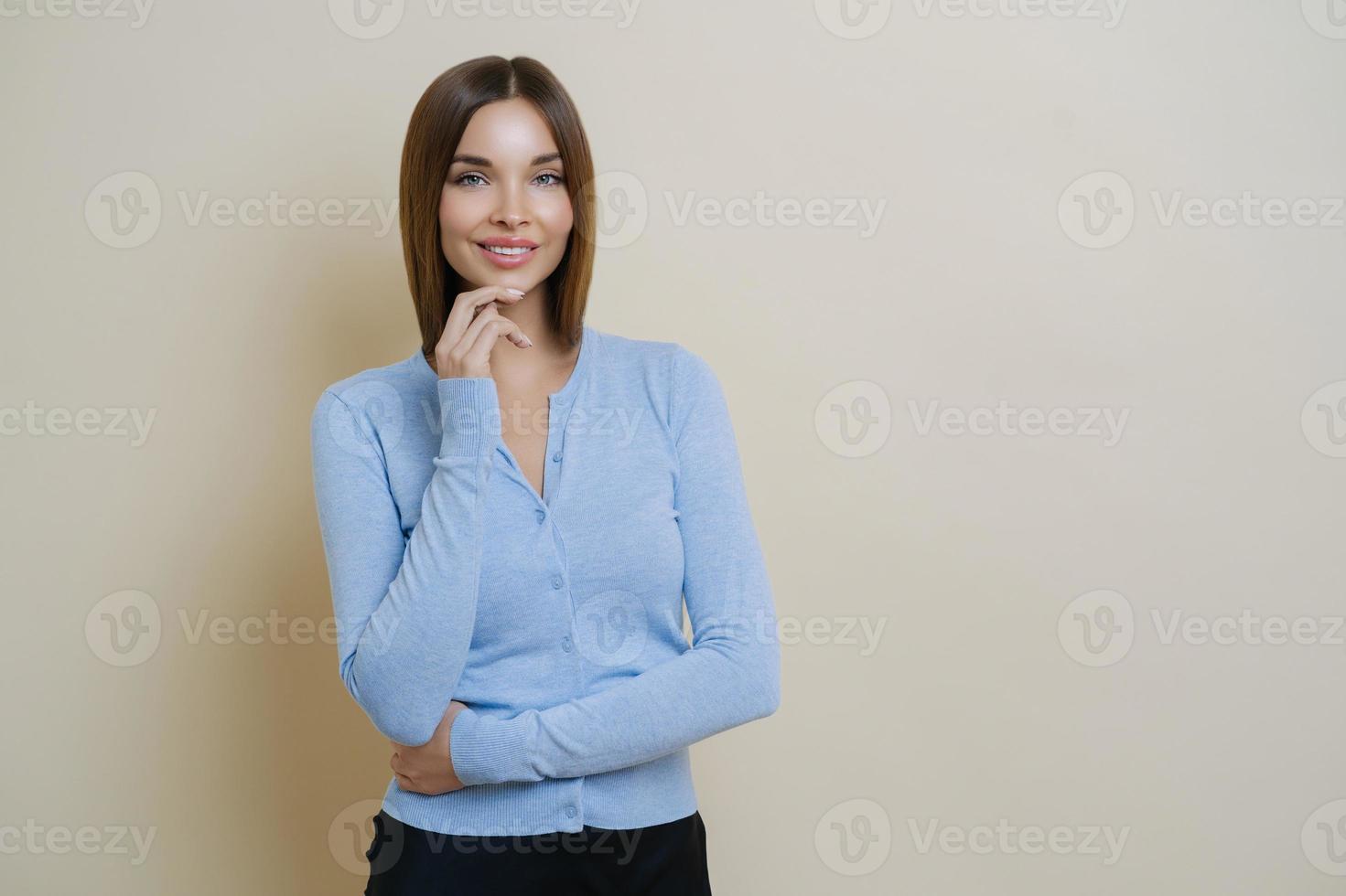 Half length shot of pretty slim woman dressed in casual blue jumper, keeps hand under chin, enjoys smooth perfect healthy skin, ready for work, isolated over beige background, blank space aside photo