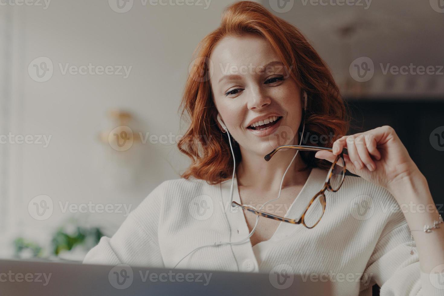 Confident pleased redhead woman concentrated in laptop computer, watches video or movie on laptop computer, enjoys friendly video call, holds transparent glasses, uses free internet connection photo