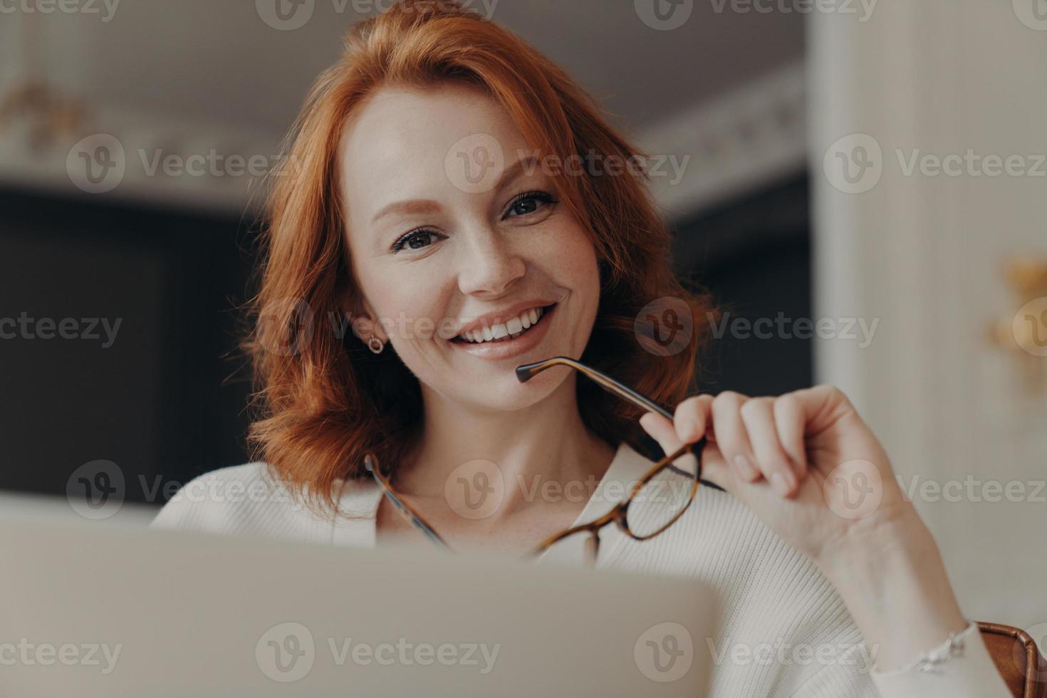 Cheerful redhead female web designer works on freelance, poses at opened laptop computer, holds glasses, shares content in social networks, smiles gently, poses at home. People and technology photo