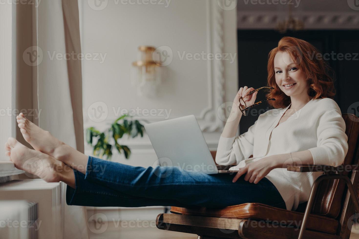 Cheerful successful ginger woman copywriter works from home, keeps laptop on knees, stretches legs, thinks on idea for creating advertising content on own website, browses information in internet photo