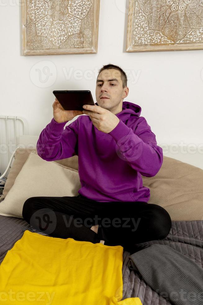 hombre con capucha violeta usando tableta digital, toma una foto de su ropa vieja para venderla en línea. venta en sitio web, comercio electrónico. reutilización, concepto de segunda mano. consumidor consciente, estilo de vida sostenible
