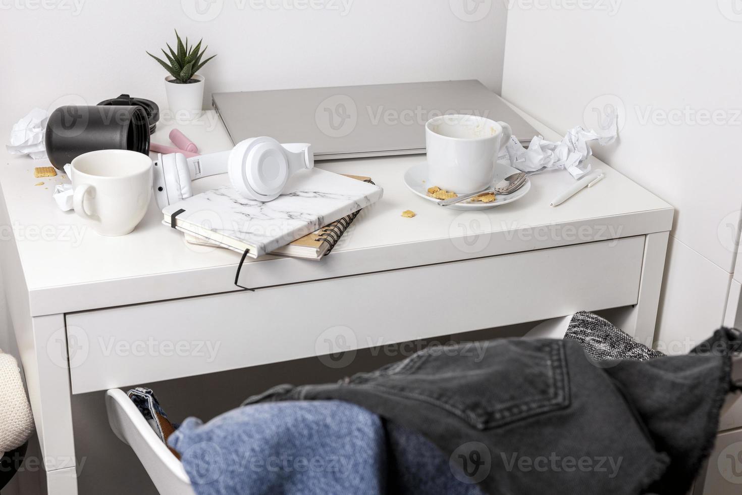 Messy table with laptop, dirty cups, and stationary items. Cluttered workplace, desk. Concept of being overwhelmed by working or studying. Mess and chaos at home. Deadline, stress. photo