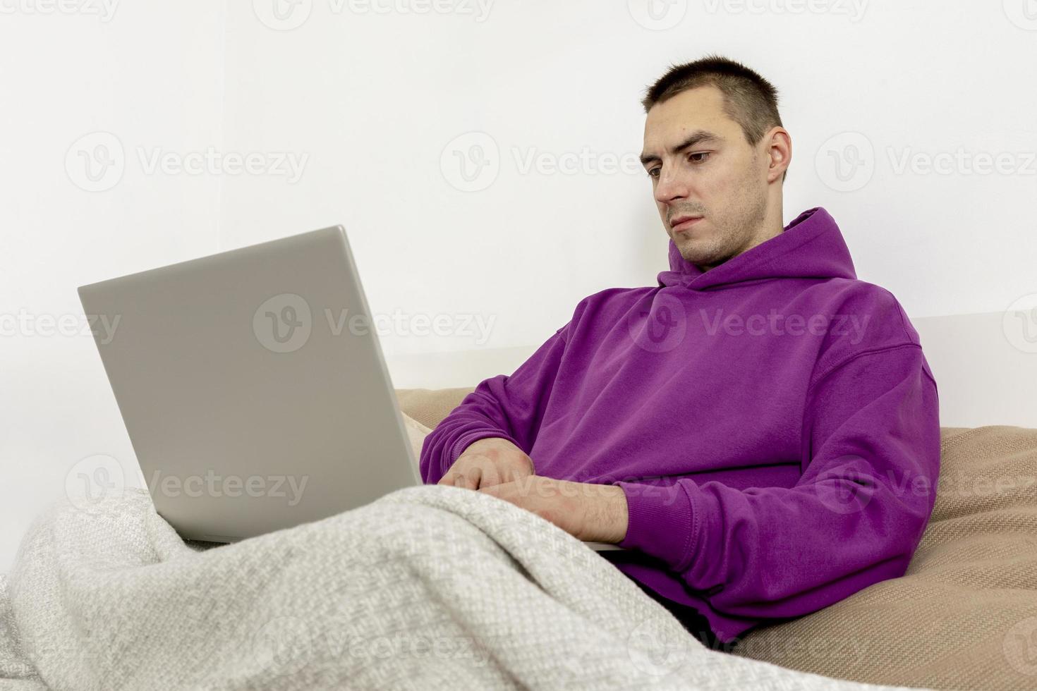 Young caucasian man with violet hoodie sitting on bed and holding laptop computer. Man using notebook to surf in internet, read news, watch movie, study or work online. Relaxing at home. photo