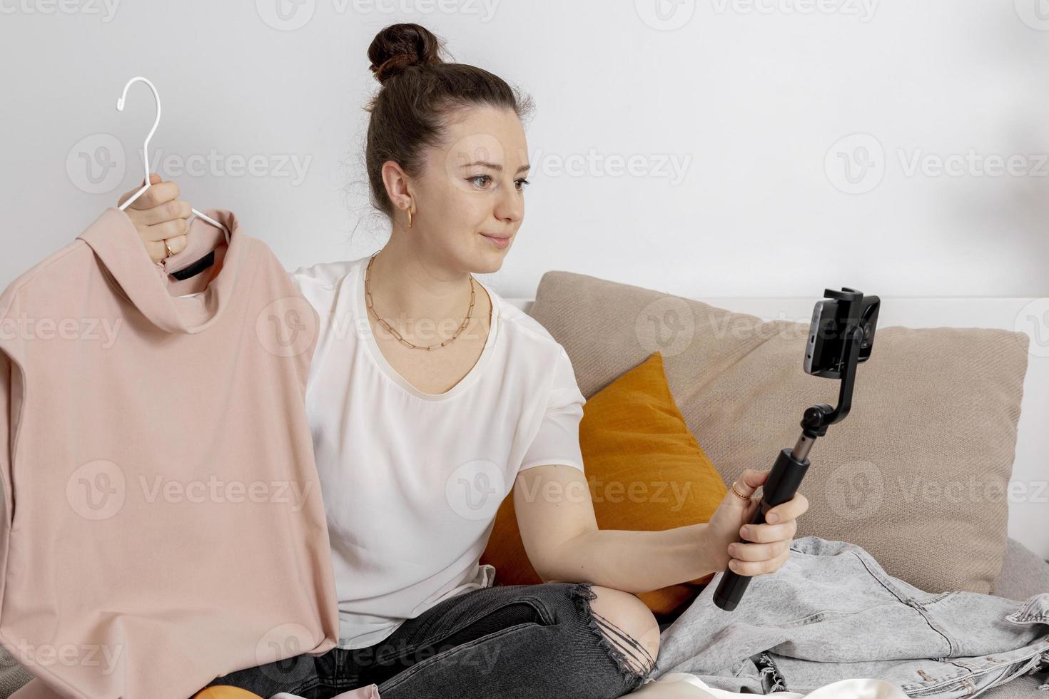 Young woman sitting on the bed at home and taking photo of her old clothes to sell them online. Selling on website, e-commerce. Reuse, second-hand concept. Conscious consumer, sustainable lifestyle.