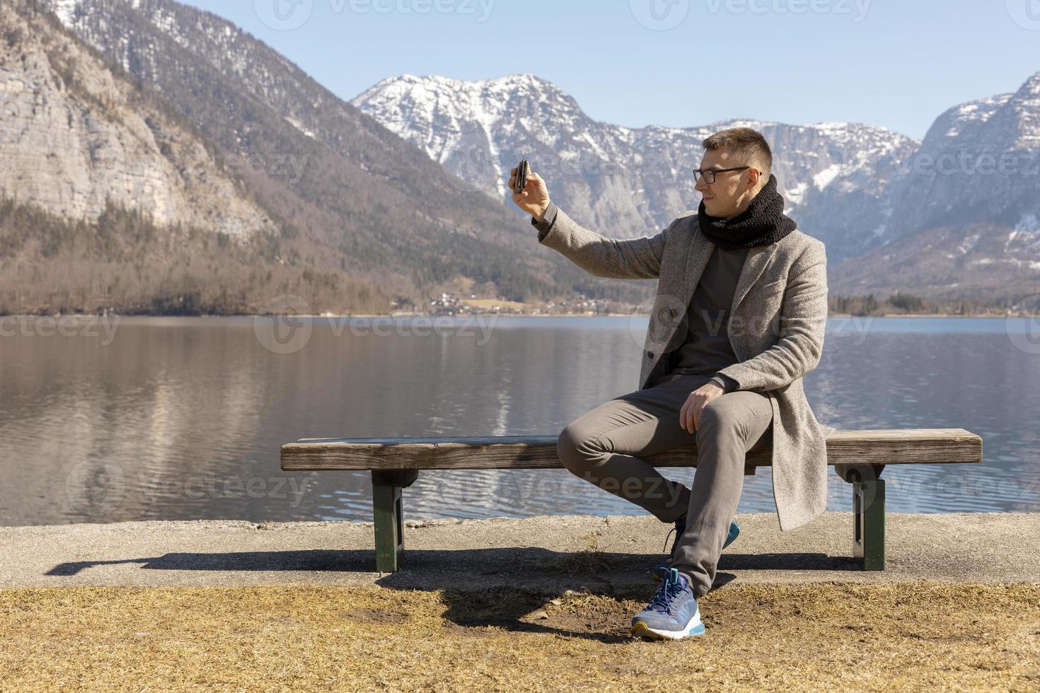 joven adulto sentado al aire libre en un banco y haciéndose selfie con su smartphone, disfrutando de las montañas, el lago, el buen tiempo, el cielo azul, el sol. hermoso, increíble paisaje. vacaciones, vacaciones, tiempo de viaje. foto