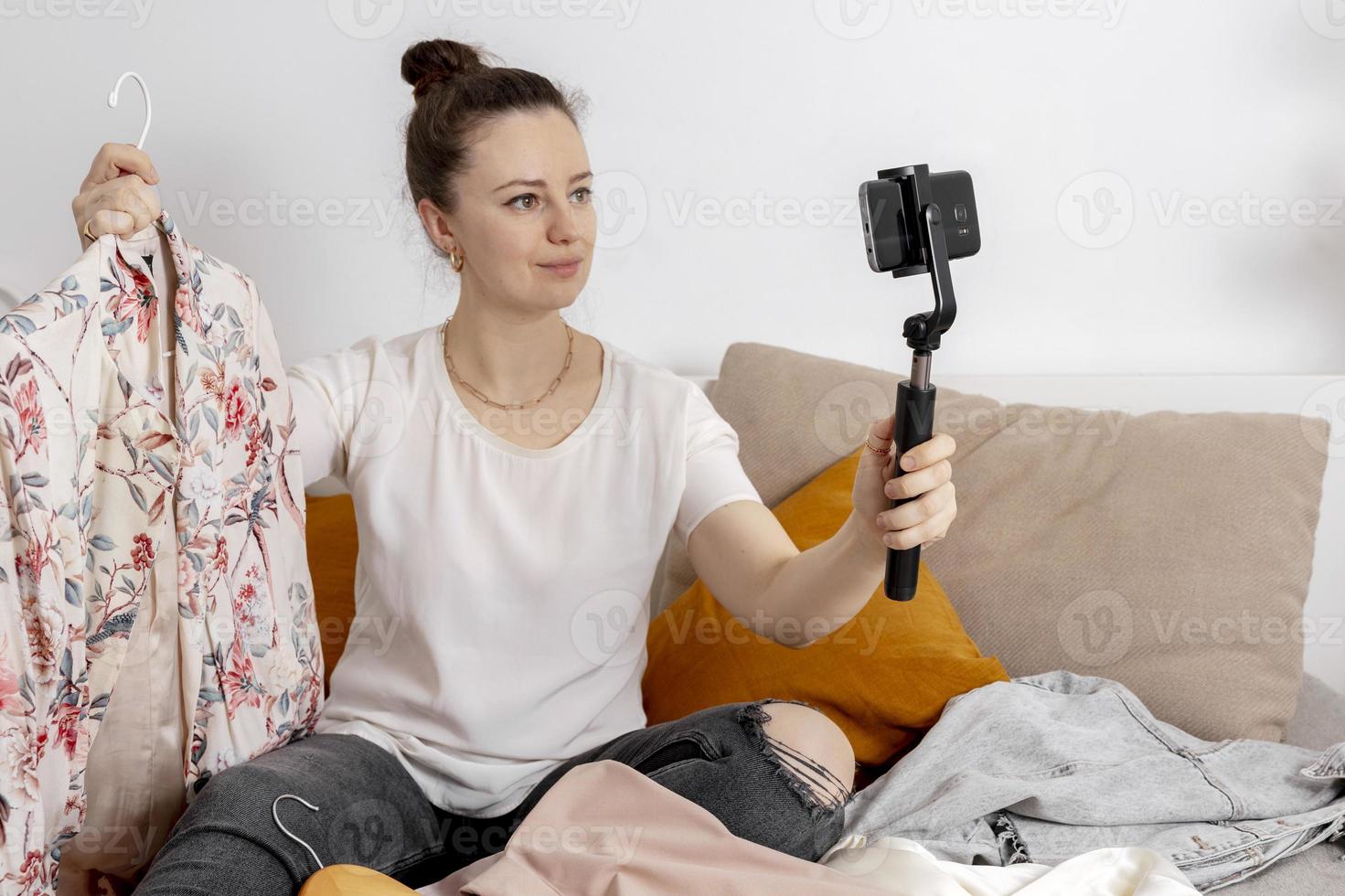 mujer joven sentada en la cama en casa y tomando fotos de su ropa vieja para venderla en línea. venta en sitio web, comercio electrónico. reutilización, concepto de segunda mano. consumidor consciente, estilo de vida sostenible.