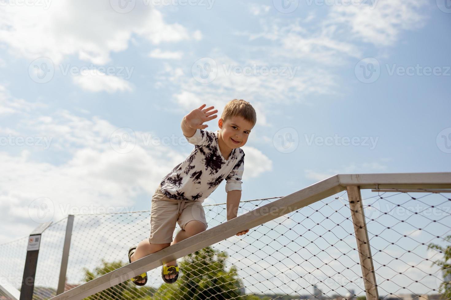 Adorable caucasian boy climbing on metal handrail outdoor. Cute child having fun in the city. Active leisure. Beautiful view, blue sky, amazing sunny weather. Go forward, climb to the top. photo