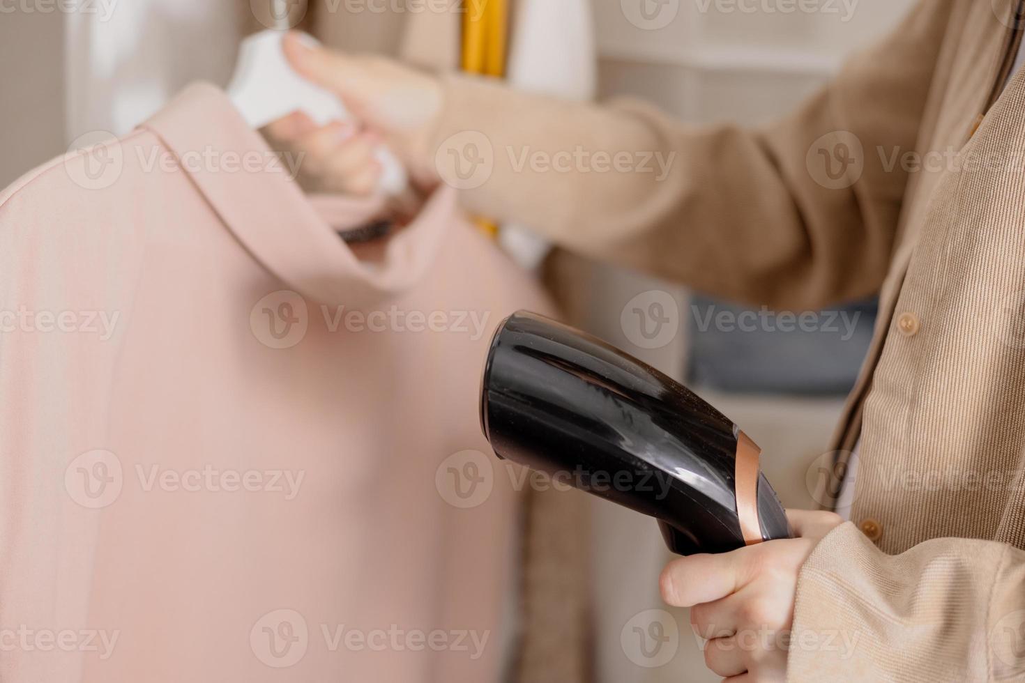 Young woman steaming her clothes at home, close up view. Female using electric steamer, ironing clothes on hanger. Modern technologies. photo