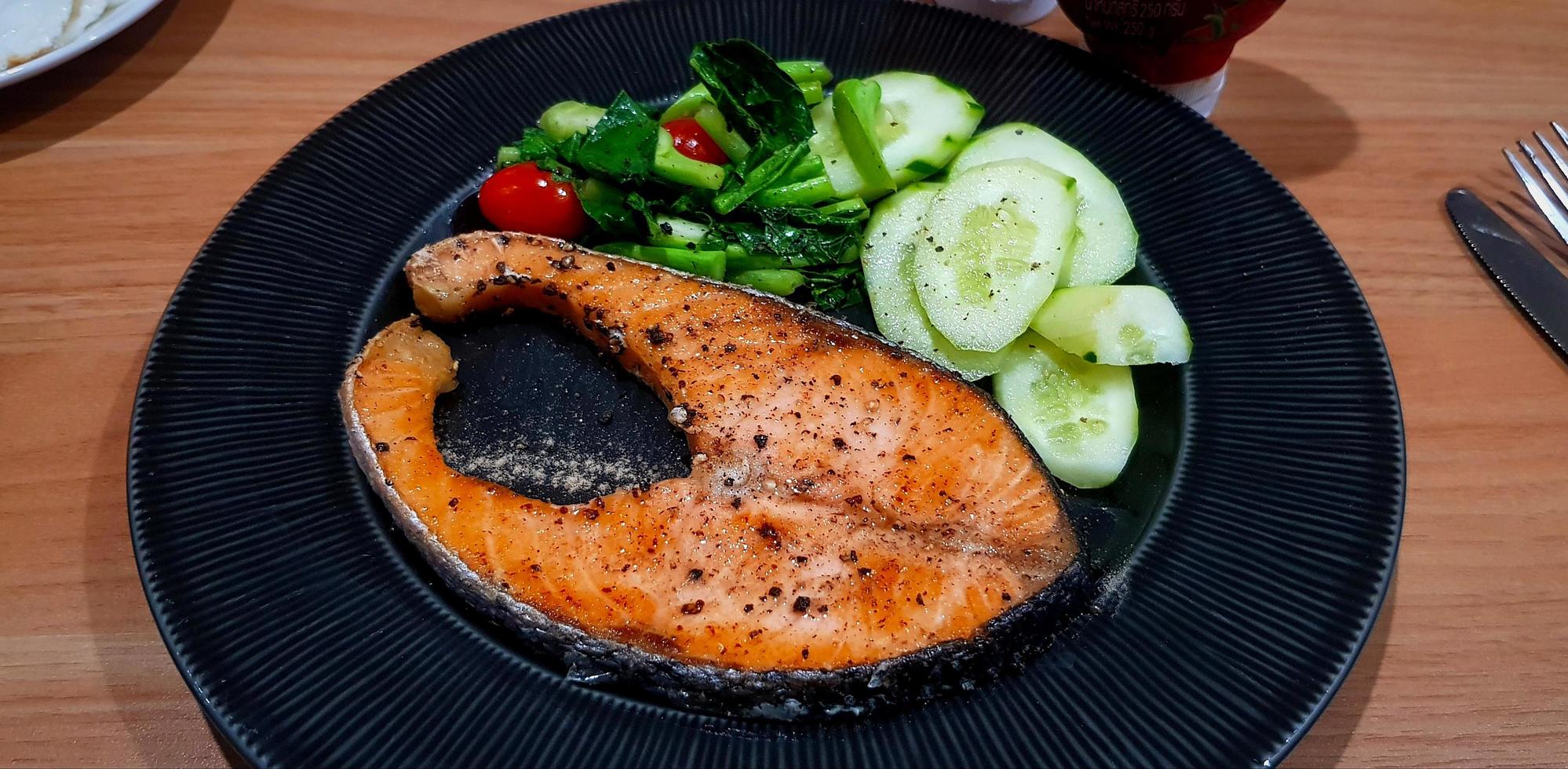 Grilled salmon steak with slices cucumber, tomato and fresh vegetable on black dish or plate on wooden table for lunch time. Healthy food and life style eating. Fish with topping black pepper and salt photo