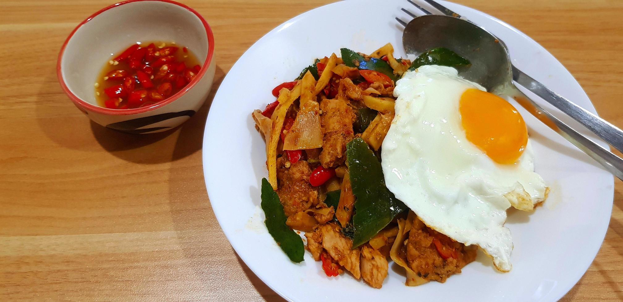 Stir fried chicken with sour bamboo shoot, red chili, kaffir lime leaves and fried egg in white plate or dish and spicy fish sauce on wooden table or wood background. Asian food with spoon and fork. photo