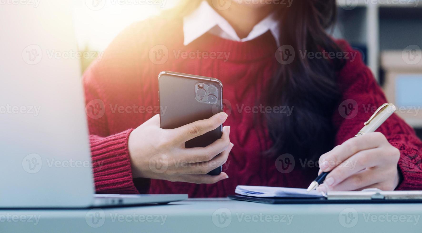 Young business woman sitting in office at table and using smartphone. On desk is laptop and tablet computer, on screen charts and graphs. Woman analyzing data. Student learning online. photo