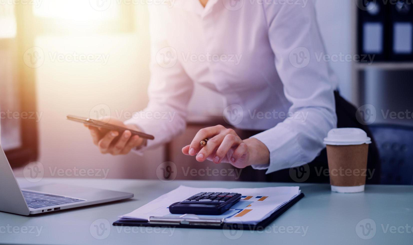 Young business woman sitting in office at table and using smartphone. On desk is laptop and tablet computer, on screen charts and graphs. Woman analyzing data. Student learning online. photo
