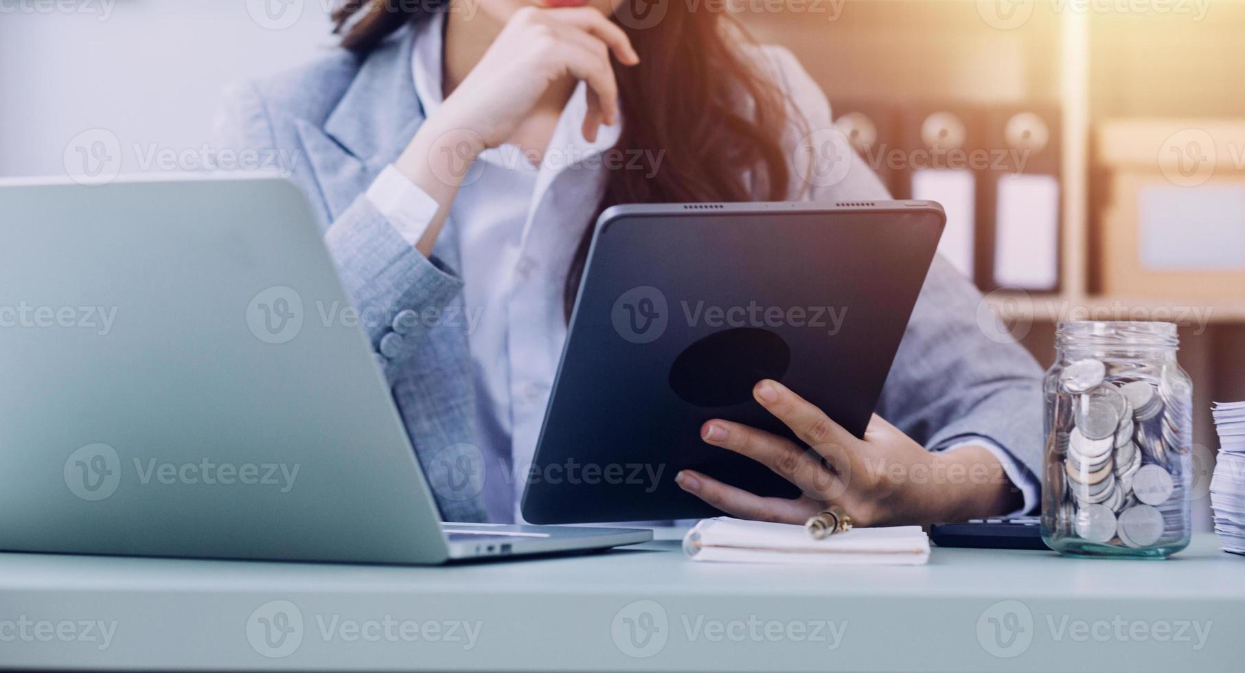 Young business woman sitting in office at table and using smartphone. On desk is laptop and tablet computer, on screen charts and graphs. Woman analyzing data. Student learning online. photo