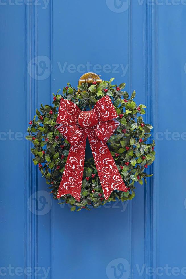 tradicional corona de navidad verde y roja colgada en la puerta de entrada azul de la casa. decoración tradicional del hogar durante la navidad foto