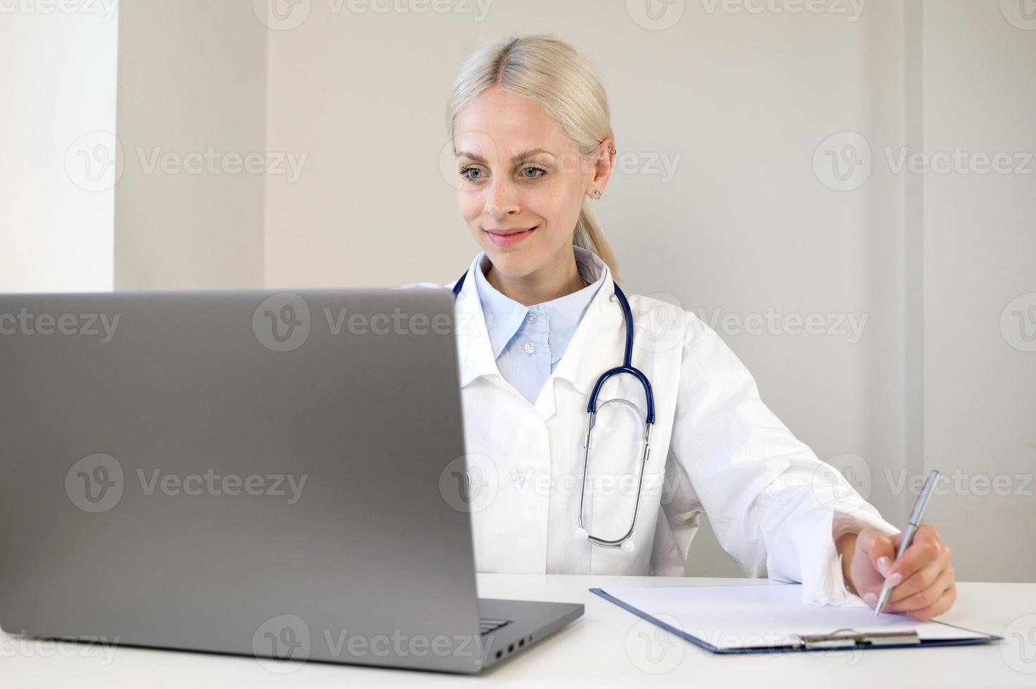 Portrait of confident young Caucasian female doctor in white medical uniform sit at desk filling out the forms looking at laptop screen photo