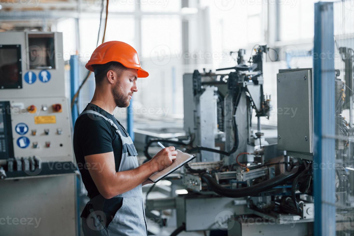 With notepad in hands. Industrial worker indoors in factory. Young technician with orange hard hat photo