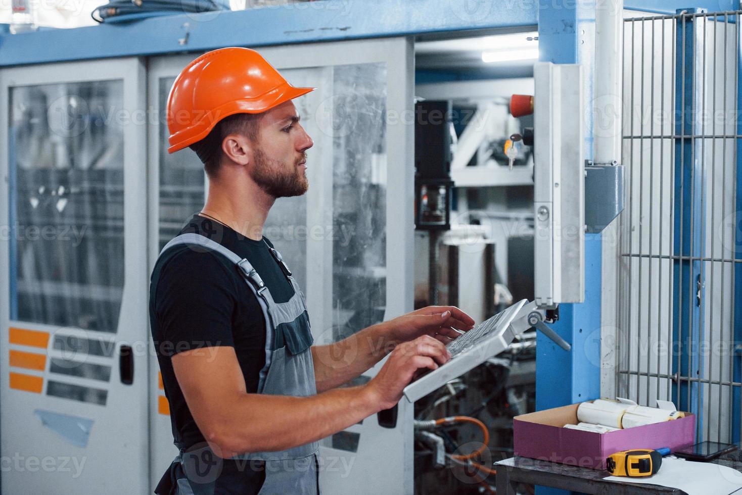 Using machine to program operations. Industrial worker indoors in factory. Young technician with orange hard hat photo