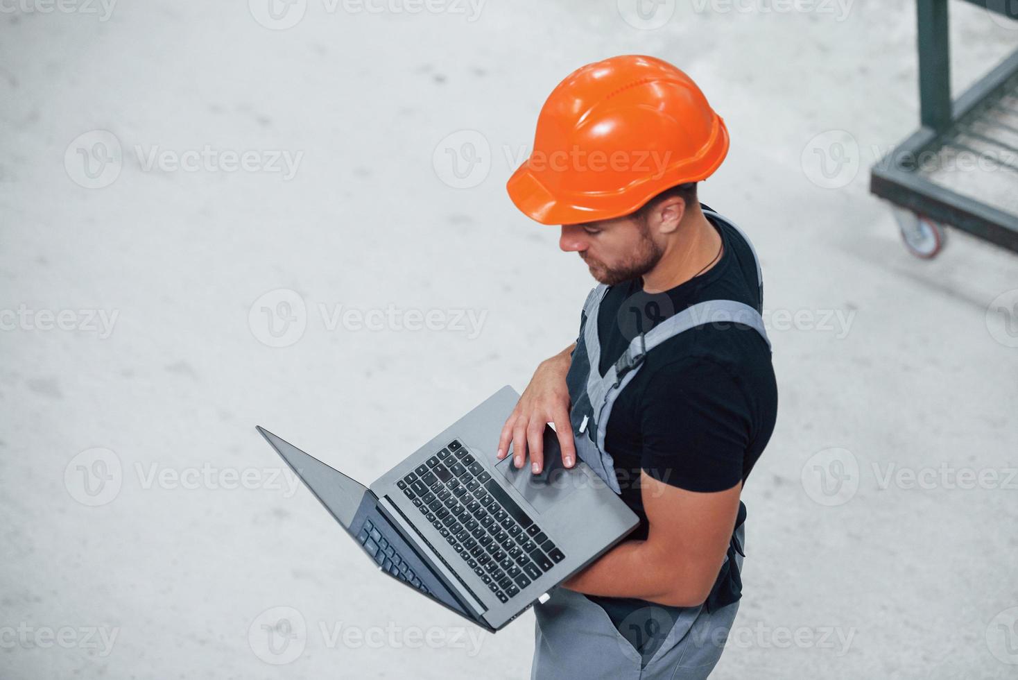 Aerial view. With laptop in hands. Industrial worker indoors in factory. Young technician with orange hard hat photo