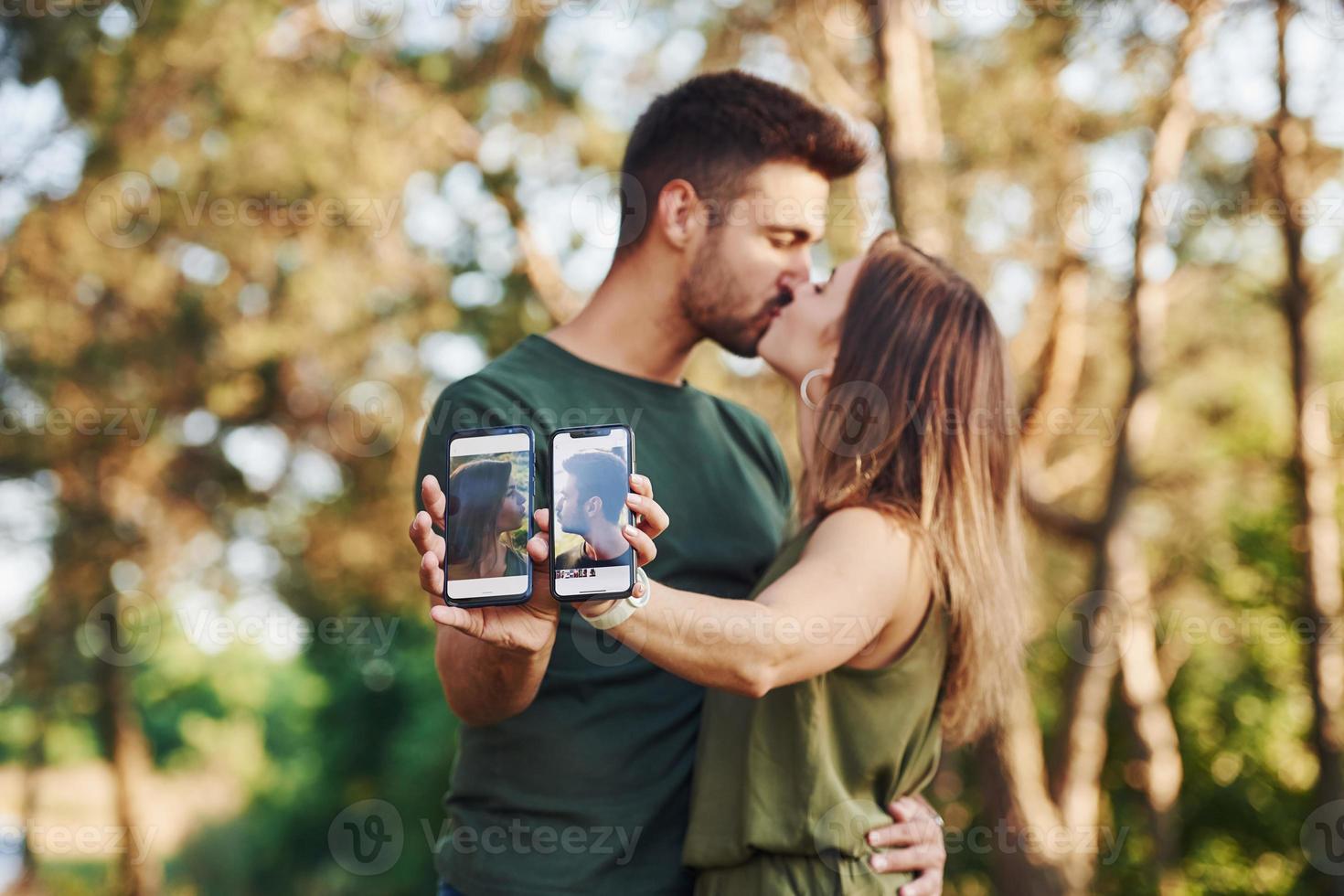 Holding two smartphones with photos of them. Beautiful young couple have a good time in the forest at daytime