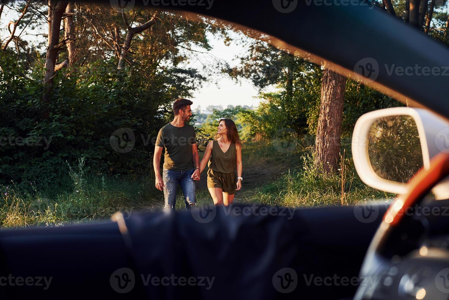 View from the car's interior. Steering wheel, side mirror. Beautiful young couple have a good time in the forest at daytime photo