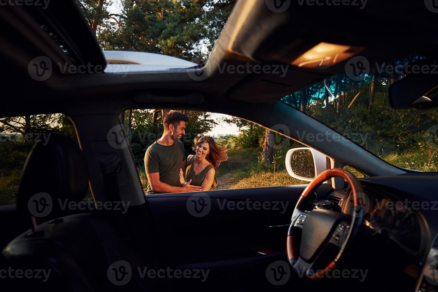 View from the car's interior. Steering wheel, side mirror. Beautiful young couple have a good time in the forest at daytime photo